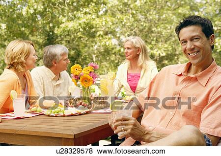 Stock Photo - Portrait of a mature man holding a glass of lemon juice with three mature people sitting behind him. Fotosearch