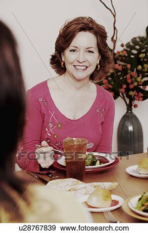 Stock Photo - Portrait of a mature woman sitting at the dining table with her friend and smiling. Fotosearch