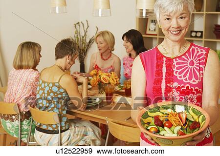 Portrait of a mature woman holding a bowl of salad and her friends sitting at the dining table in the background View Large Photo Image Stock Photo - Portrait of a mature woman holding a bowl of salad and her friends sitting at the dining table in the background. Fotosearch