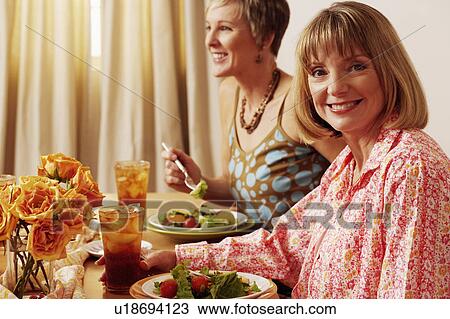 Stock Image - Portrait of a mature woman sitting at the dining table with her friend and smiling. Fotosearch