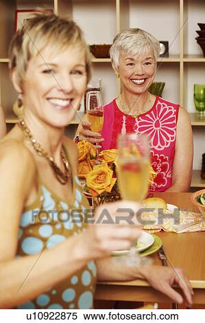 Stock Photography - Portrait of a mature woman sitting at the dining table with her friend and holding a champagne flute. Fotosearch