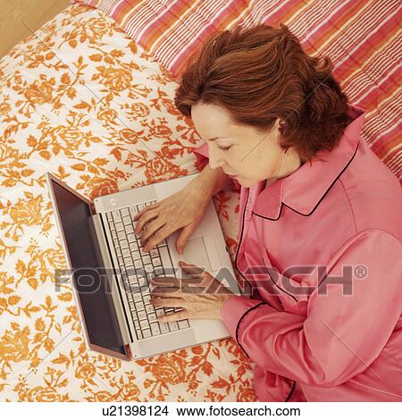 Picture - High angle view of a mature woman lying in bed using a laptop. Fotosearch
