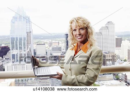 Stock Photo - Portrait of a businesswoman holding a laptop with buildings in the background. Fotosearch