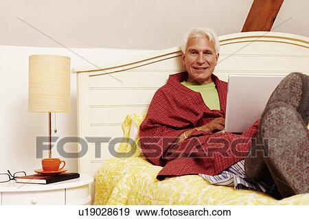 Portrait of a mature man leaning on the bed with a laptop View Large Photo Image Stock Photo - Portrait of a mature man leaning on the bed with a laptop. Fotosearch