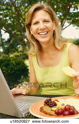 Stock Photo - Portrait of a mature woman using a laptop and eating fruit salad. Fotosearch
