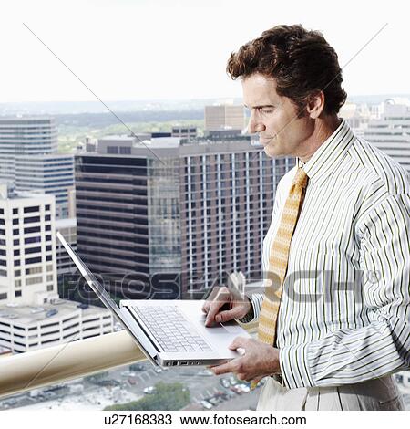 Stock Image - Side profile of a businessman using a laptop in a balcony with a cityscape in the background. Fotosearch
