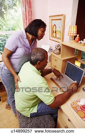 Stock Photo - Side profile of a mature man using a laptop with a mature woman standing near him. Fotosearch