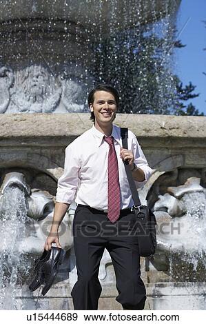 Businessman standing in front of fountain with laptop bag and shoes in hand smiling. View Large Photo Image Stock Photo - Businessman standing in front of fountain with laptop bag and shoes in hand smiling.. Fotosearch
