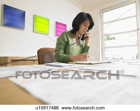 Stock Photograph - Businesswoman writing and giving a call at her desk. Low angle.. Fotosearch