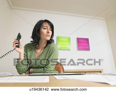 Stock Image - Businesswoman holding a phone at her desk.. Fotosearch