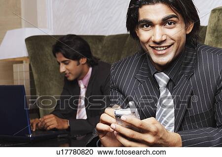 Stock Image - Portrait of a businessman holding a mobile phone with another businessman using a laptop behind him. Fotosearch
