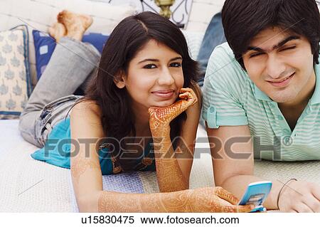 Stock Photography - Portrait of a young woman holding a mobile phone with a young man lying beside her on the bed. Fotosearch