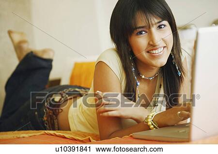 Stock Image - Portrait of a young woman lying on the bed in front of a laptop. Fotosearch