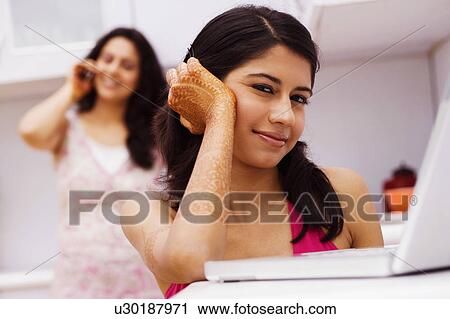Stock Image - Portrait of a young woman sitting in front of a laptop with a mid adult woman talking on a mobile phone in the background. Fotosearch