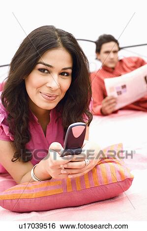 Young woman holding a mobile phone and a young man reading a newspaper in the background View Large Photo Image Stock Photograph - Young woman holding a mobile phone and a young man reading a newspaper in the background. Fotosearch