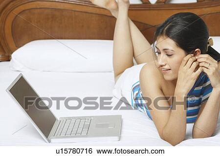 Young woman lying on the bed and looking at a laptop View Large Photo Image Stock Photograph - Young woman lying on the bed and looking at a laptop. Fotosearch