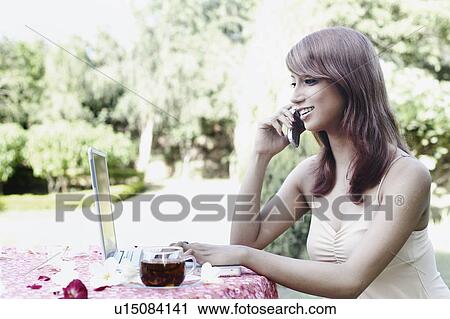 Stock Image - Young woman talking on a mobile phone and using a laptop. Fotosearch