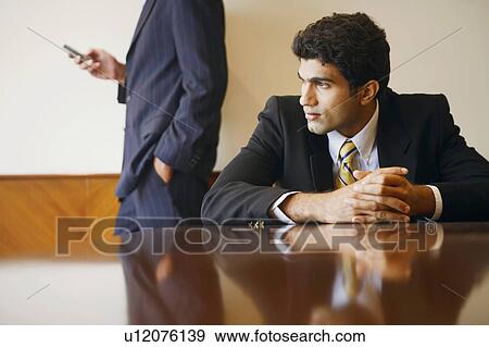 Close-up of a businessman sitting at a table with another holding a mobile phone behind him View Large Photo Image Stock Photo - Close-up of a businessman sitting at a table with another holding a mobile phone behind him. Fotosearch