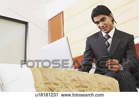Stock Image - Portrait of a businessman holding a credit card and sitting in front of a laptop. Fotosearch