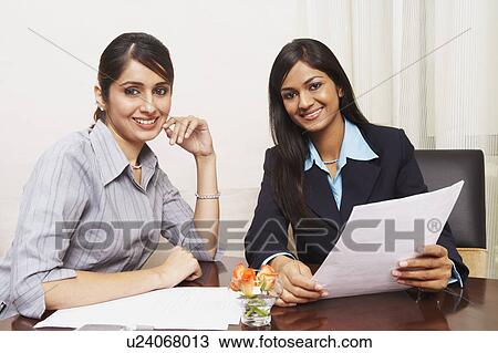 Stock Image - Portrait of two businesswomen sitting in an office and smiling. Fotosearch