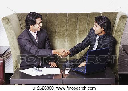 Stock Image - Two businessmen sitting on a couch and shaking hands. Fotosearch
