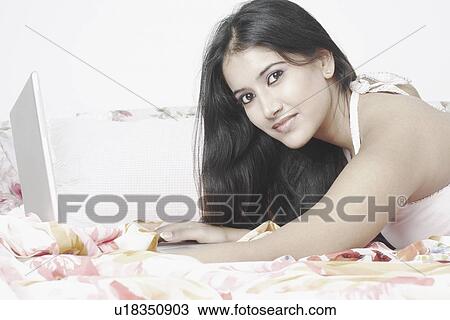 Stock Image - Portrait of a young woman lying on the bed using a laptop. Fotosearch