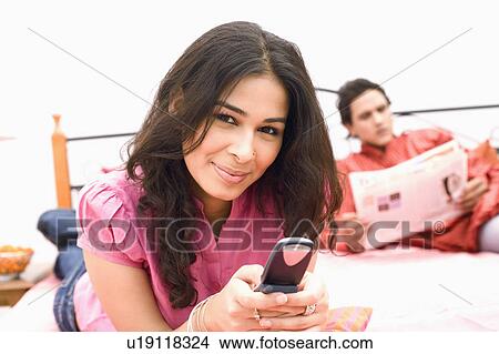 Picture - Young woman holding a mobile phone and a young man reading a newspaper in the background. Fotosearch