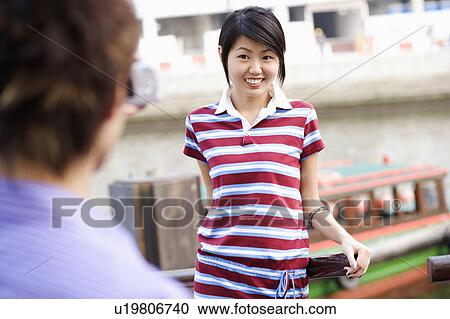Stock Image - Rear view of a young man taking a picture of a young woman. Fotosearch
