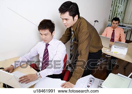 Stock Image - Businessman using a laptop with another businessman standing behind him. Fotosearch