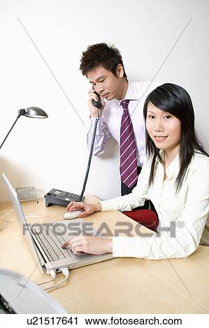 Stock Image - Businesswoman using a laptop with a businessman talking on the telephone. Fotosearch