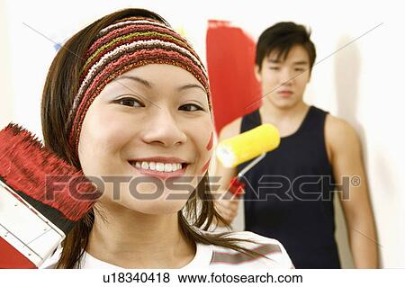 Stock Photo - Portrait of a young woman holding a paint brush and a young man standing in the background. Fotosearch