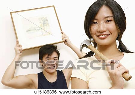 Portrait of a young woman holding a hammer with a young man standing in the background holding a picture frame View Large Photo Image Stock Photograph - Portrait of a young woman holding a hammer with a young man standing in the background holding a picture frame. Fotosearch