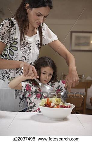 Stock Photo - Woman helping daughter (5-7) to toss salad. Fotosearch