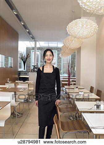 Stock Image - Young waitress standing in empty restaurant, smiling, portrait. Fotosearch