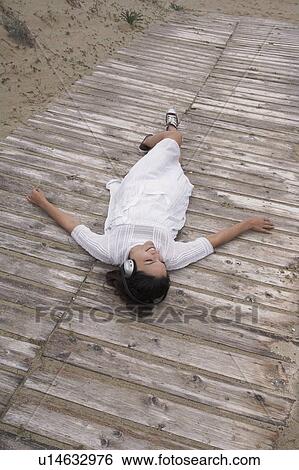 Stock Photograph - Woman wearing headphones lying on boardwalk. Fotosearch