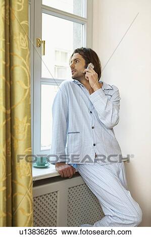 Stock Photography - young man wearing pyjamas looking out of window holding mobile phone. Fotosearch