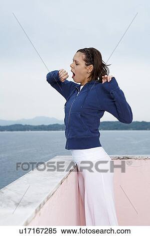 Stock Photography - Young woman on balcony yawning morning sea and sky in background.. Fotosearch