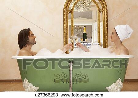 Stock Image - young couple sitting in bath toasting glasses of champagne, side view. Fotosearch