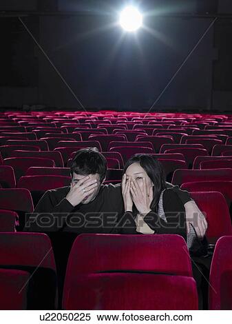 Stock Photography - Young couple sitting in cinema holding hands over faces. Fotosearch