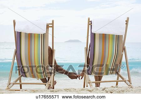 Stock Image - Couple on lounge chairs, holding hands (rear view), St. John, US Virgin Islands, USA. Fotosearch