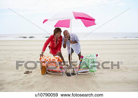 Stock Photo - Couple pointing and laughing under beach umbrella. Fotosearch