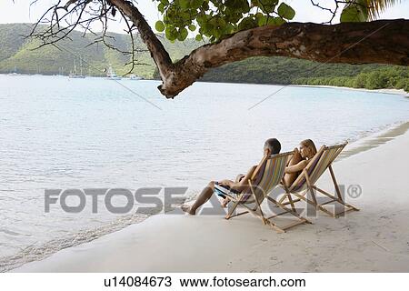 Stock Image - Mid adult couple on lounge chairs on beach (high angle view), St. John, US Virgin Islands, USA. Fotosearch
