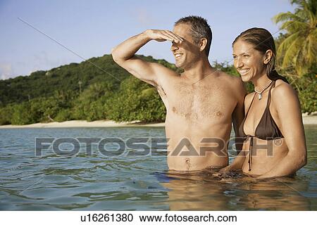 Stock Image - Mid adult couple standing in water and looking at view, St. John, US Virgin Islands, USA. Fotosearch