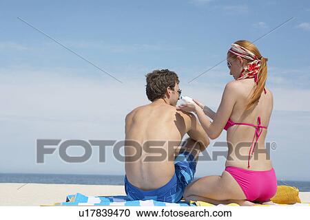 Stock Image - Woman applying sunscreen on man's back at beach. Fotosearch