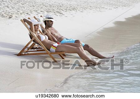 Stock Image - Young couple on lounge chairs on sandy beach. Fotosearch