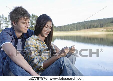 Stock Photograph - Teenage couple using mobile phone by lake. Fotosearch