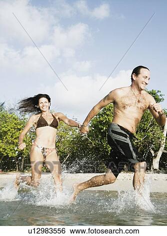 Stock Photograph - Mid adult couple running in water on the beach. Fotosearch