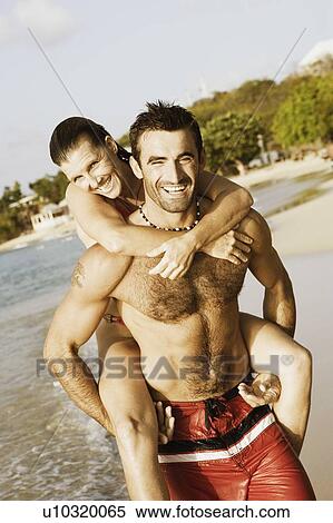 Stock Photography - Mid adult man riding piggyback on a mid adult woman on the beach. Fotosearch