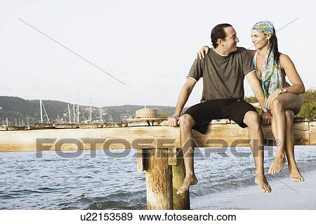 Mid adult woman with her arm around a mid adult man sitting on a jetty View Large Photo Image Stock Photo - Mid adult woman with her arm around a mid adult man sitting on a jetty. Fotosearch