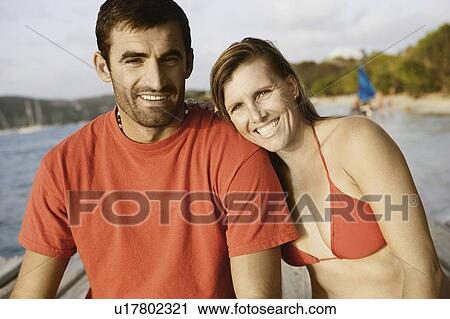 Stock Image - Portrait of a mid adult couple sitting on a pier and smiling. Fotosearch